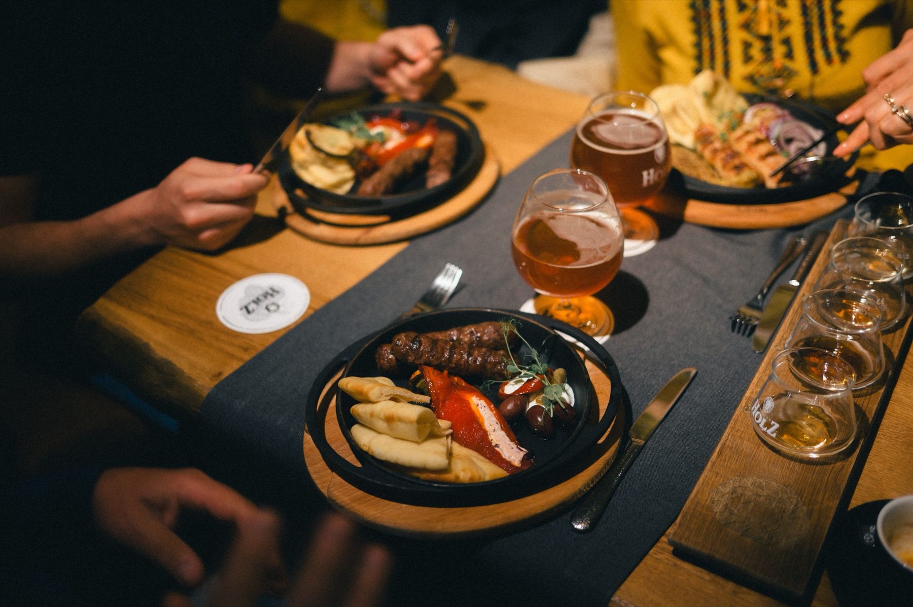 Gasten aan een houten tafel met gedeelde gerechten en glazen bier in warm avondlicht