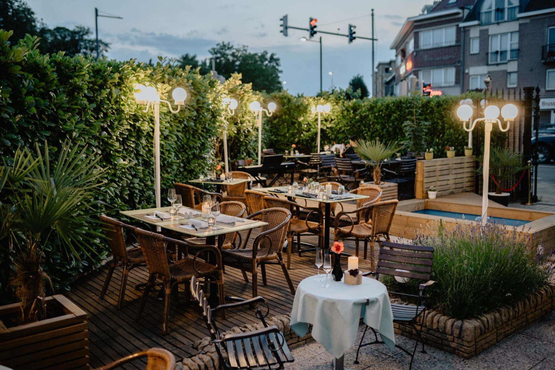Terras in de avond met gedekte tafels, rieten stoelen, kaarsjes en bolle lantaarns tussen de hagen